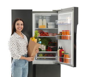 Young woman with bag of groceries near open refrigerator on white background