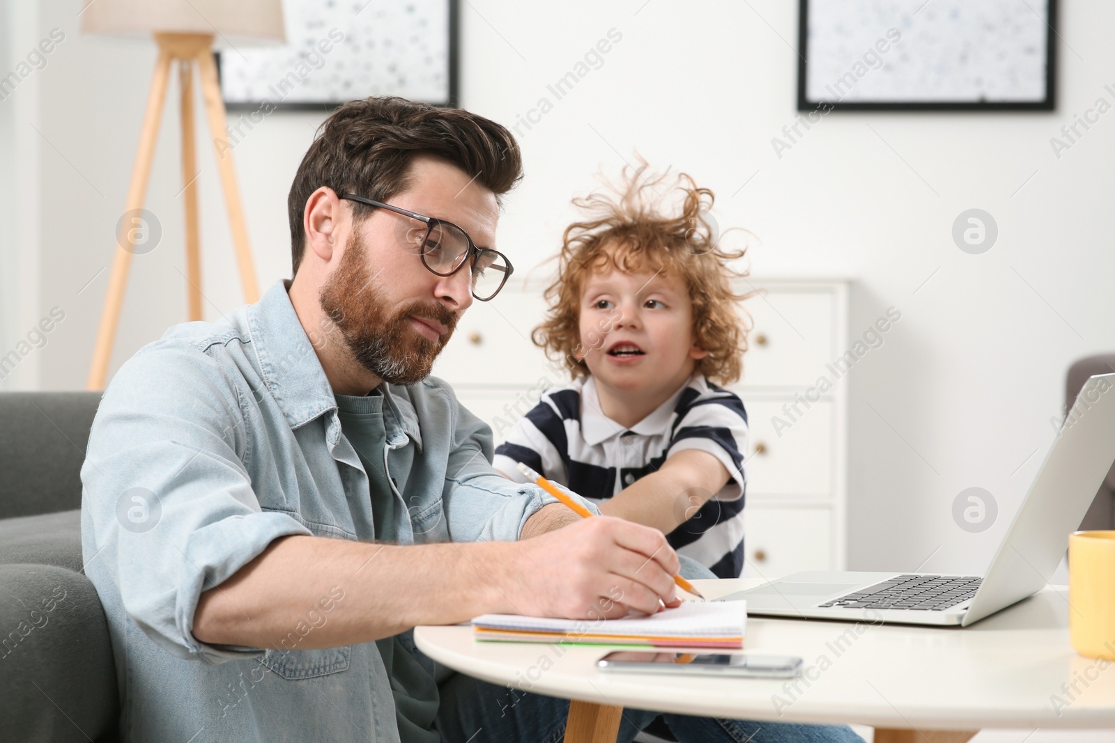 Photo of Little boy bothering his father at home. Man working remotely at desk