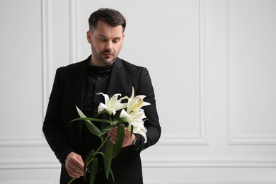 Photo of Sad man with lily flowers near white wall, space for text. Funeral ceremony