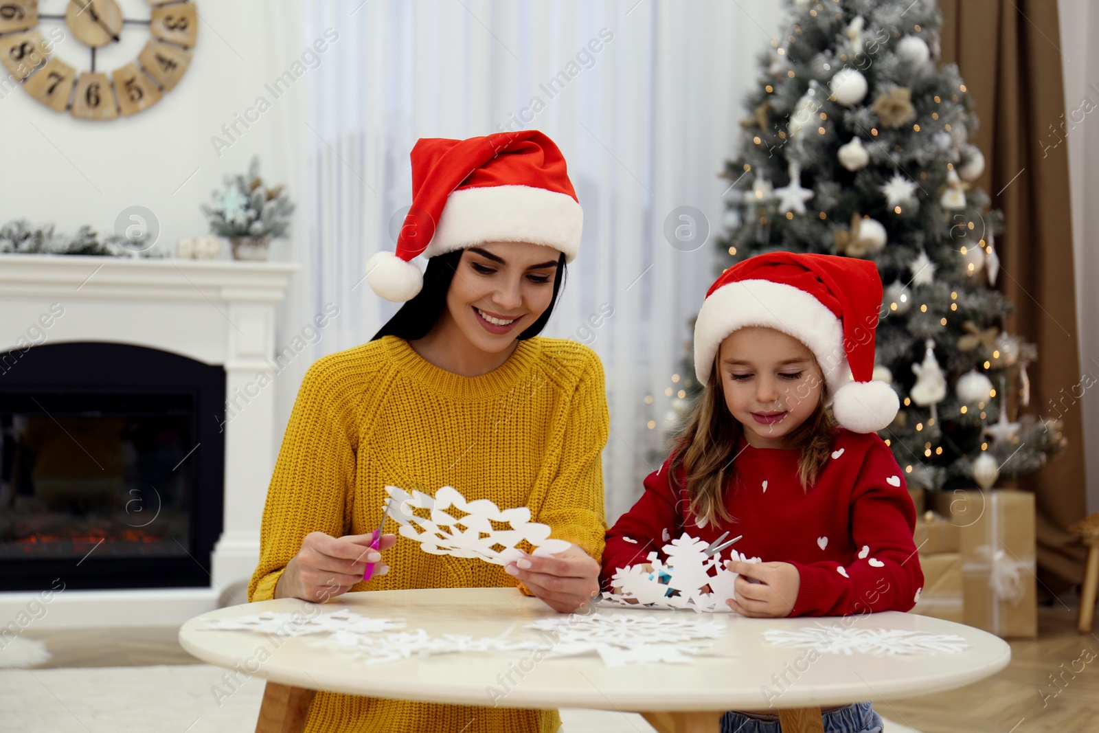 Photo of Mother and daughter in Santa hats making paper snowflakes near Christmas tree at home