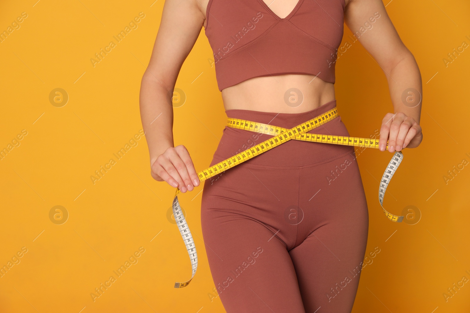 Photo of Woman in sportswear measuring waist with tape on yellow background, closeup. Space for text