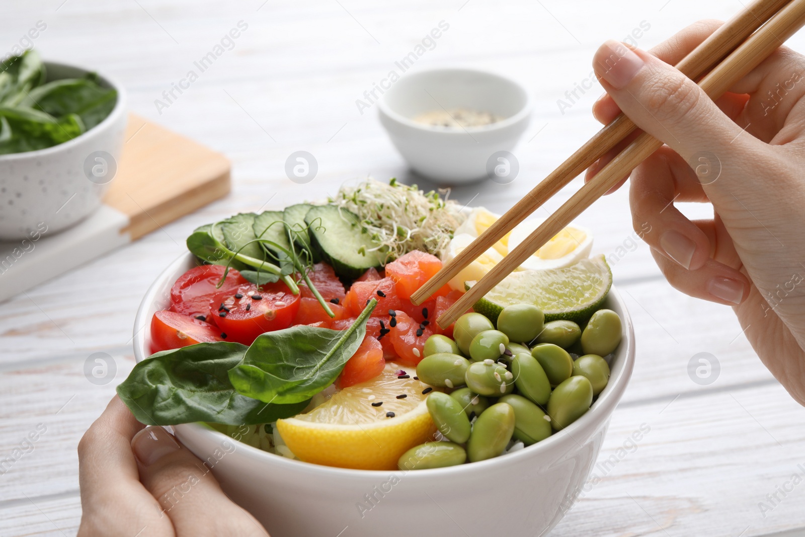 Photo of Woman eating delicious poke bowl with quail eggs, fish and edamame beans at white wooden table, closeup