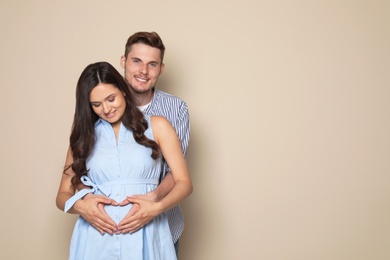 Photo of Young husband and his pregnant wife showing heart with hands on color background. Space for text