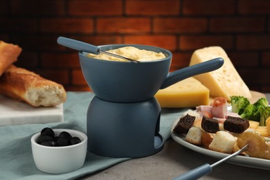 Photo of Tasty melted cheese in fondue pot, fork and snacks on grey table, closeup