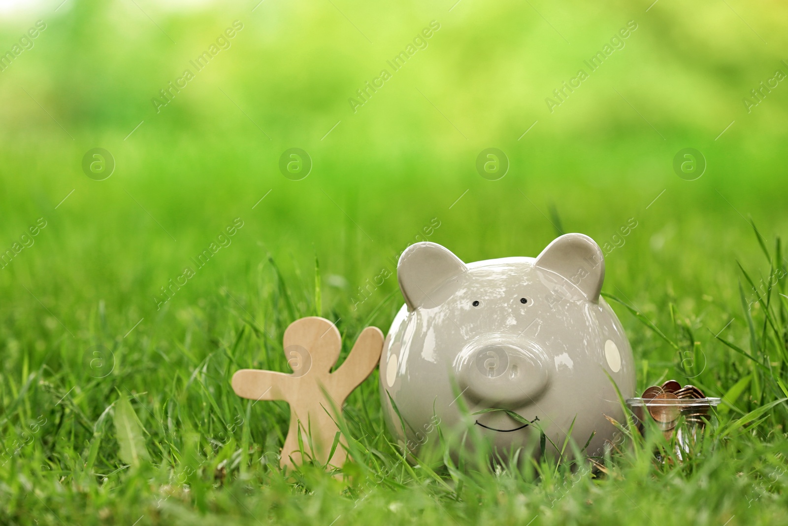 Photo of Piggy bank, wooden person and jar with coins on green grass in park. Space for text