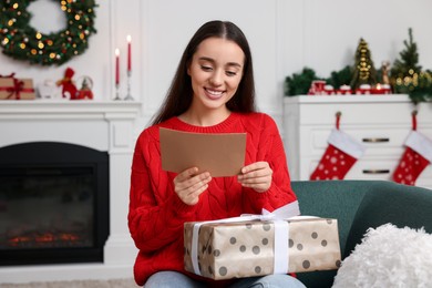 Photo of Happy young woman with Christmas gift reading greeting card at home