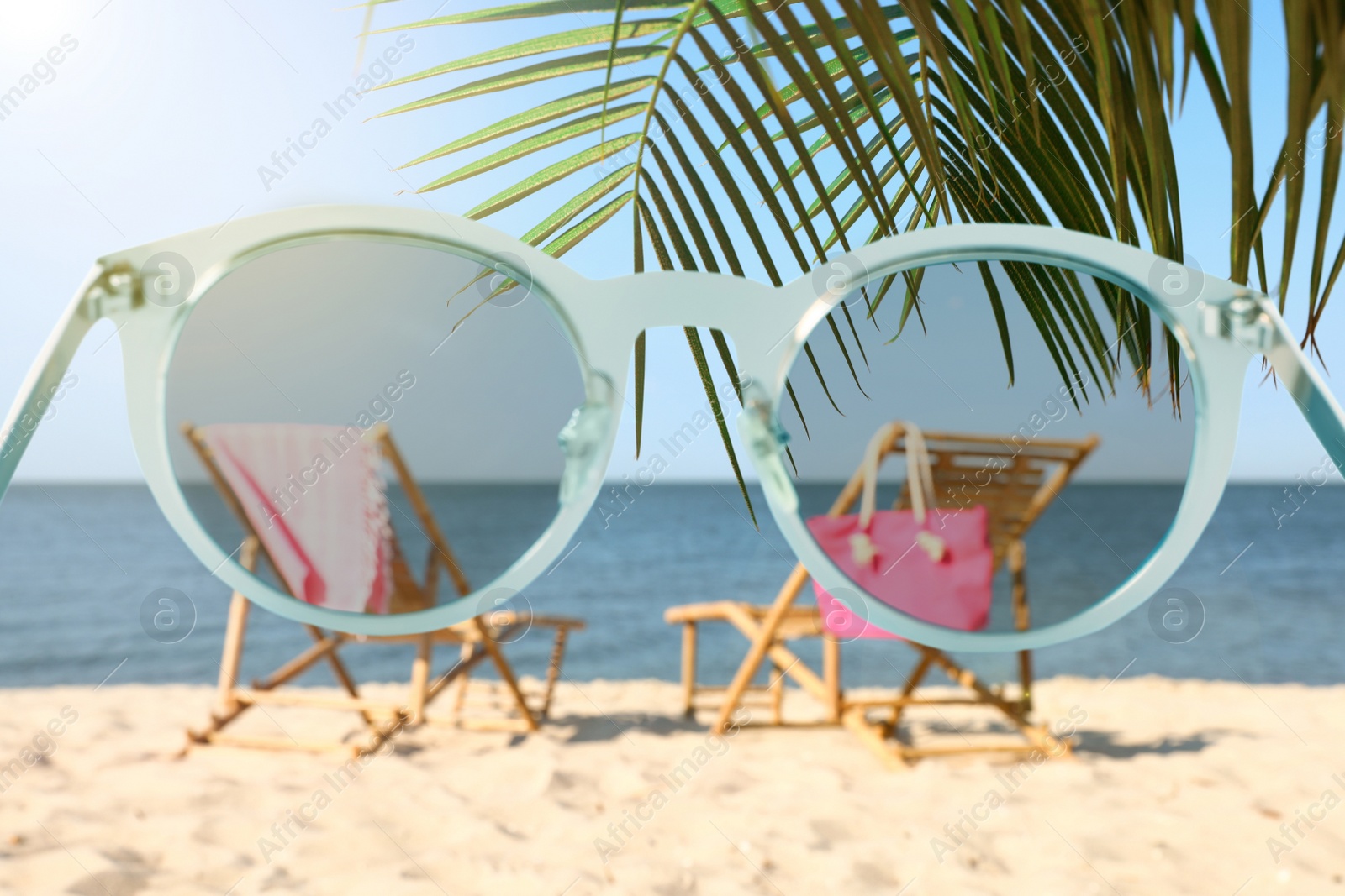 Image of Wooden sunbeds and beach accessories on sandy shore, blurred view through sunglasses