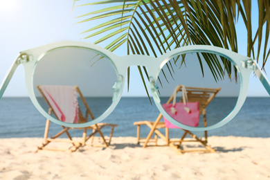 Image of Wooden sunbeds and beach accessories on sandy shore, blurred view through sunglasses