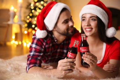 MYKOLAIV, UKRAINE - JANUARY 27, 2021: Young couple holding bottles of Coca-Cola in room decorated for Christmas, focus on hands