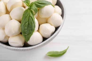 Tasty mozzarella balls and basil leaves in bowl on white wooden table, closeup. Space for text