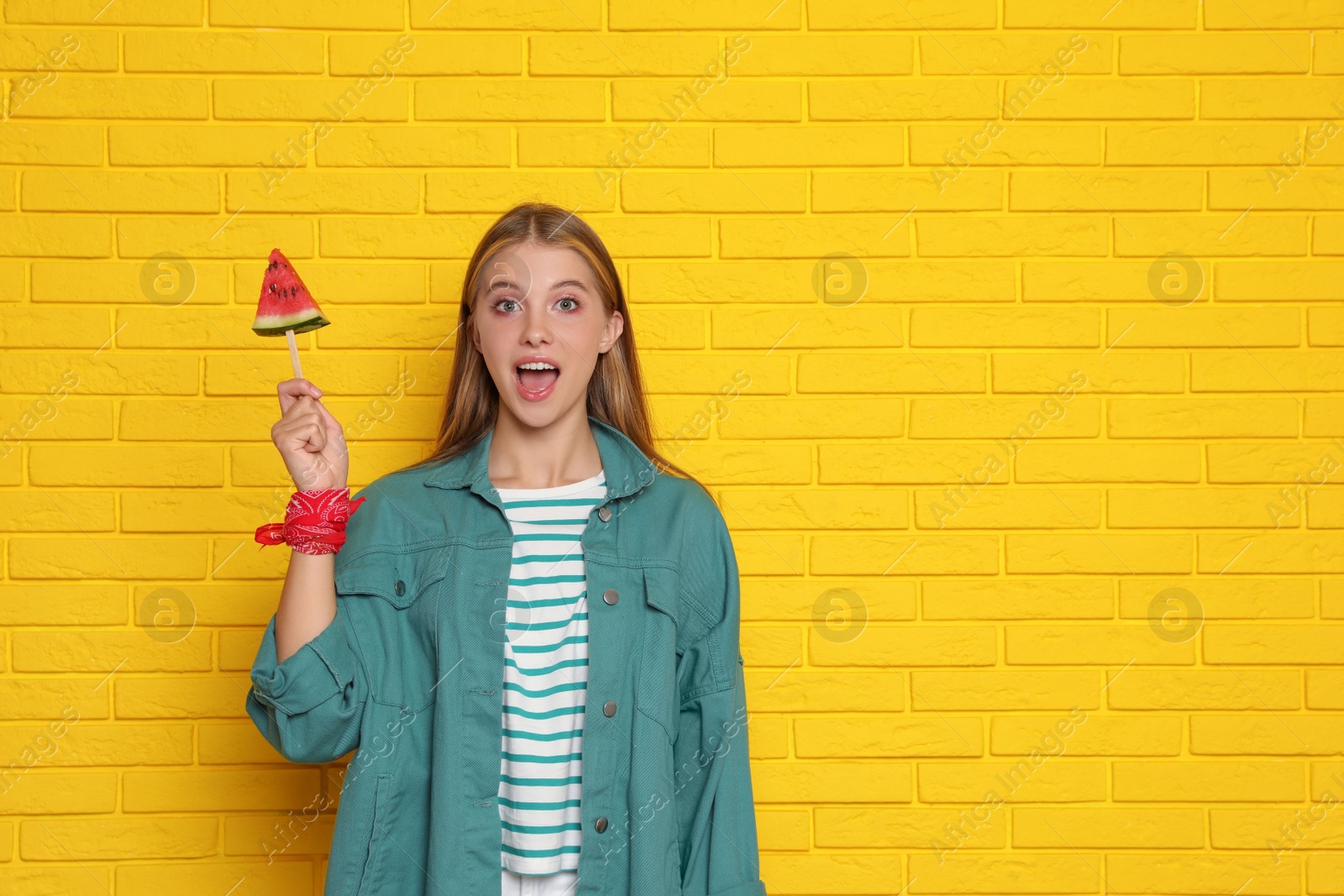 Photo of Beautiful girl with piece of watermelon near yellow brick wall. Space for text