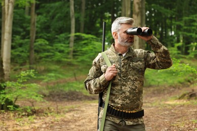 Man with hunting rifle looking through binoculars in forest. Space for text