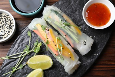 Photo of Tasty spring rolls, sauce, lime, sesame seeds and microgreens on wooden table, top view