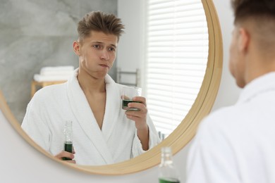 Young man using mouthwash near mirror in bathroom