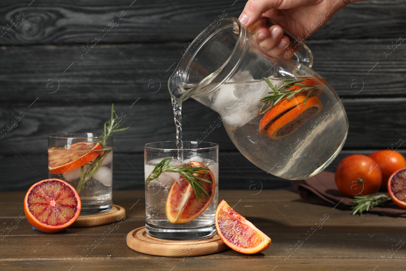 Photo of Woman pouring refreshing drink with sicilian orange from jug into glass at wooden table, closeup