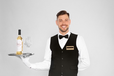 Handsome waiter holding tray with glasses and bottle of wine on light background