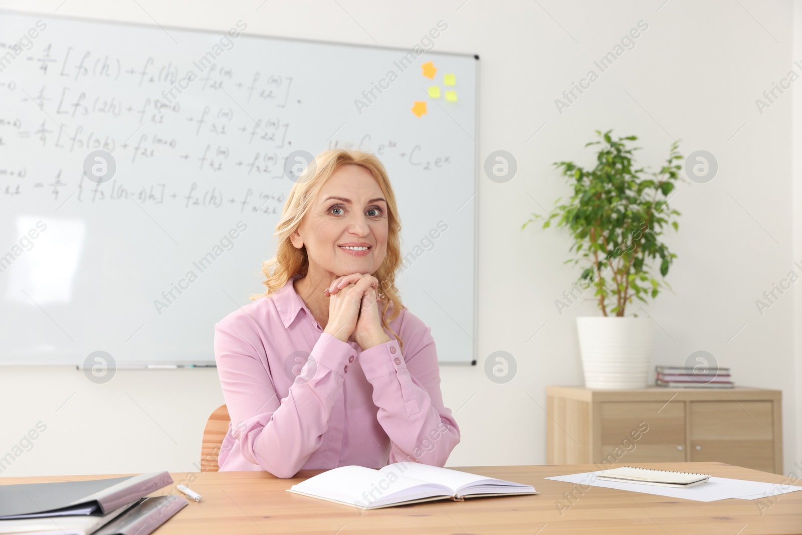 Photo of Happy teacher with book sitting at desk in classroom