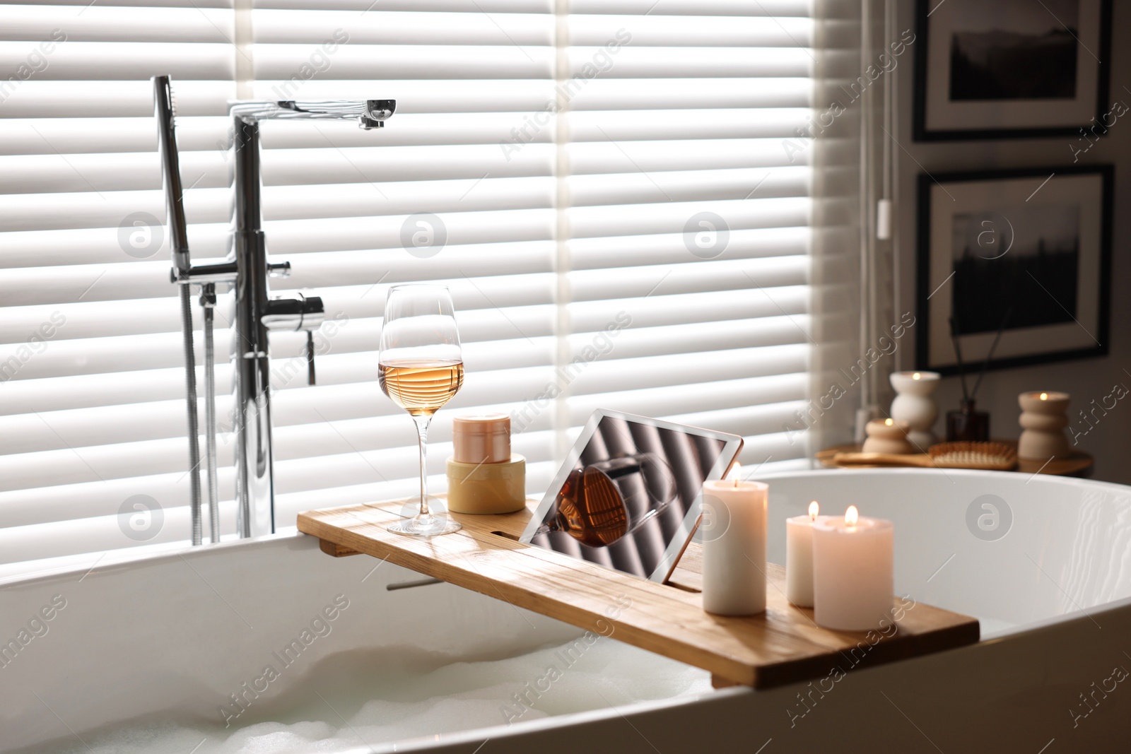 Photo of Wooden tray with tablet, wine and candles on bathtub in bathroom