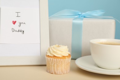 Happy Father's Day. Tasty cupcake, cup, card with phrase I Love You, Daddy in frame and gift box on beige table, closeup