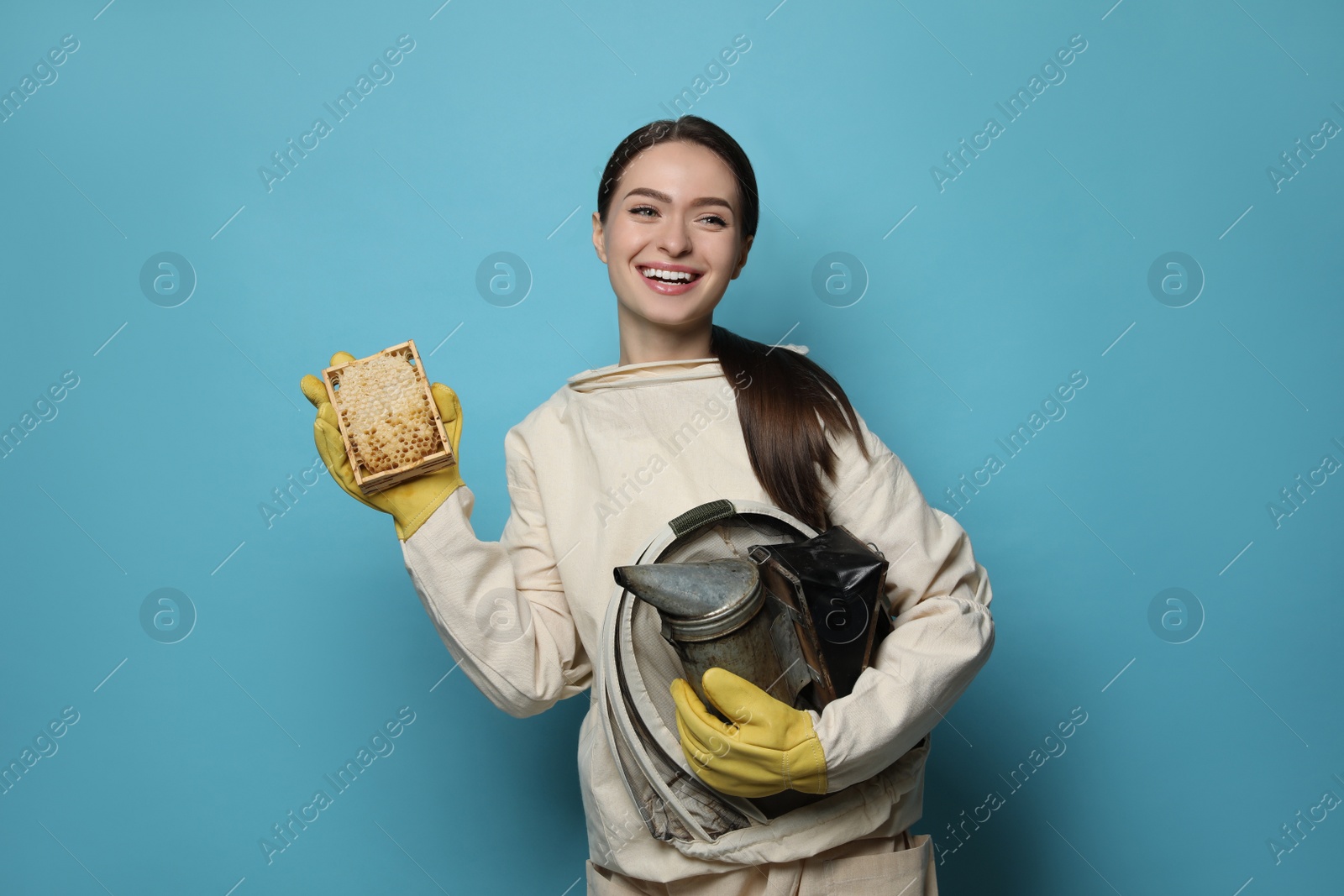 Photo of Beekeeper in uniform holding smokepot and hive frame with honeycomb on light blue background