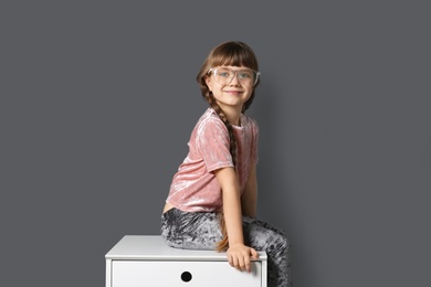 Cute little girl sitting on chest of drawers unit near gray wall