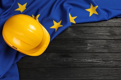 Photo of Yellow hard hat and flag of European Union on black wooden table, flat lay. Space for text