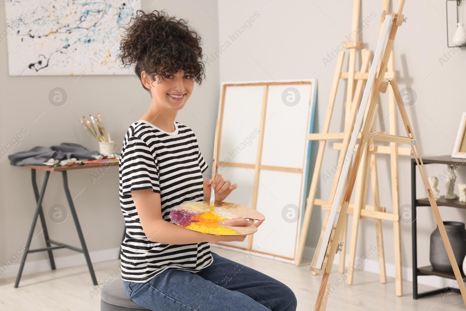 Photo of Young woman mixing paints on palette with brush near easel in studio