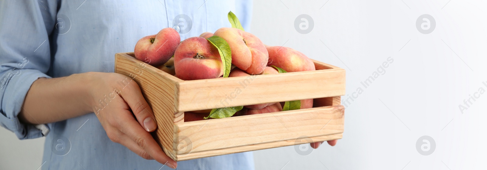 Image of Woman holding wooden crate with fresh flat peaches on light background, closeup. Banner design with space for text