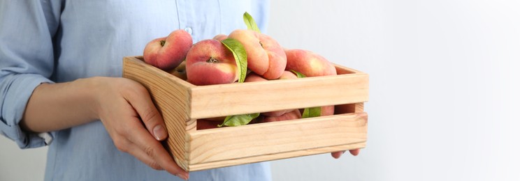 Image of Woman holding wooden crate with fresh flat peaches on light background, closeup. Banner design with space for text