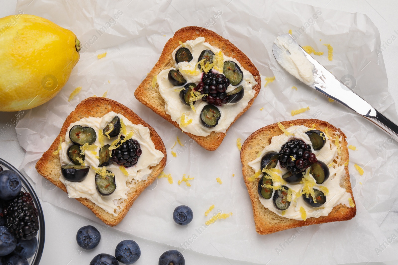 Photo of Tasty sandwiches with cream cheese, blueberries, blackberries and lemon zest on white table, flat lay