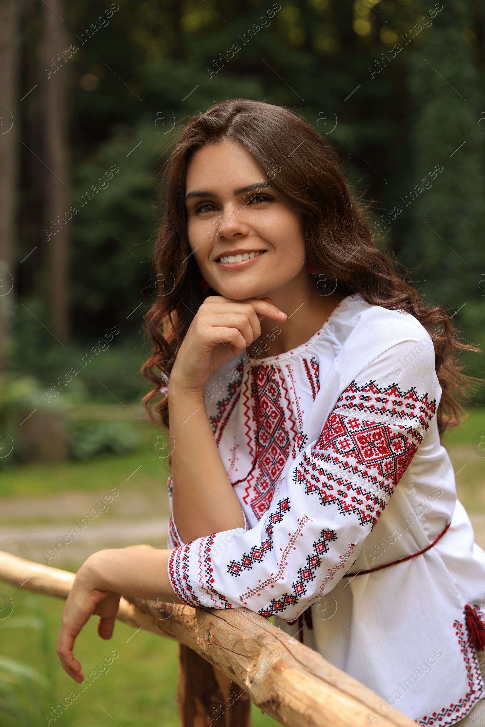 Photo of Beautiful woman wearing embroidered shirt near wooden railing in countryside. Ukrainian national clothes