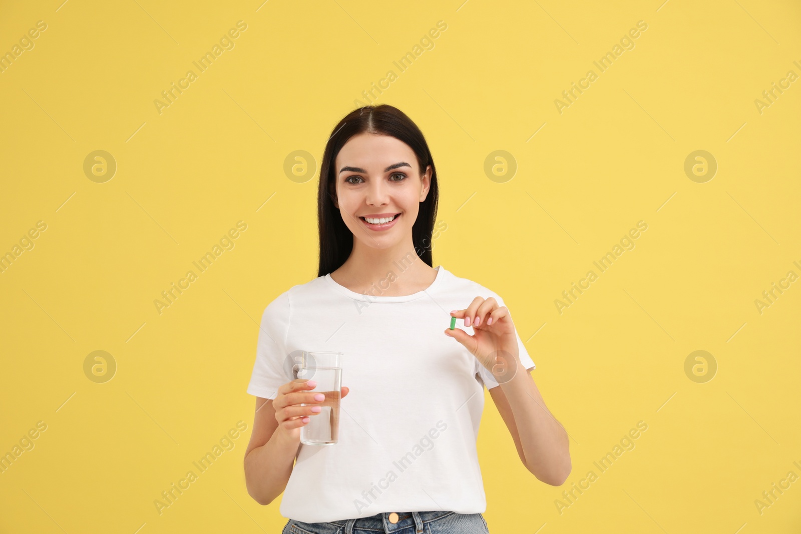 Photo of Young woman with glass of water and vitamin capsule on yellow background