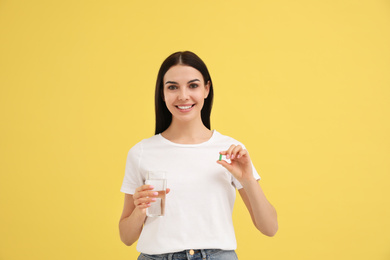 Young woman with glass of water and vitamin capsule on yellow background