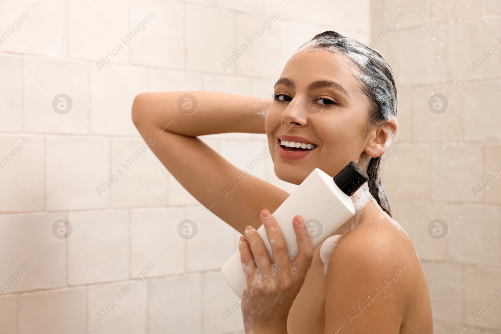 Photo of Happy woman with bottle of shampoo in shower at home. Washing hair