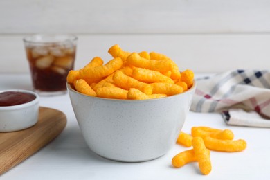 Crunchy cheesy corn snack in bowl, ketchup and refreshing drink on white wooden table, closeup