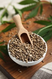 Photo of Bowl with hemp seeds and scoop on wooden board, closeup