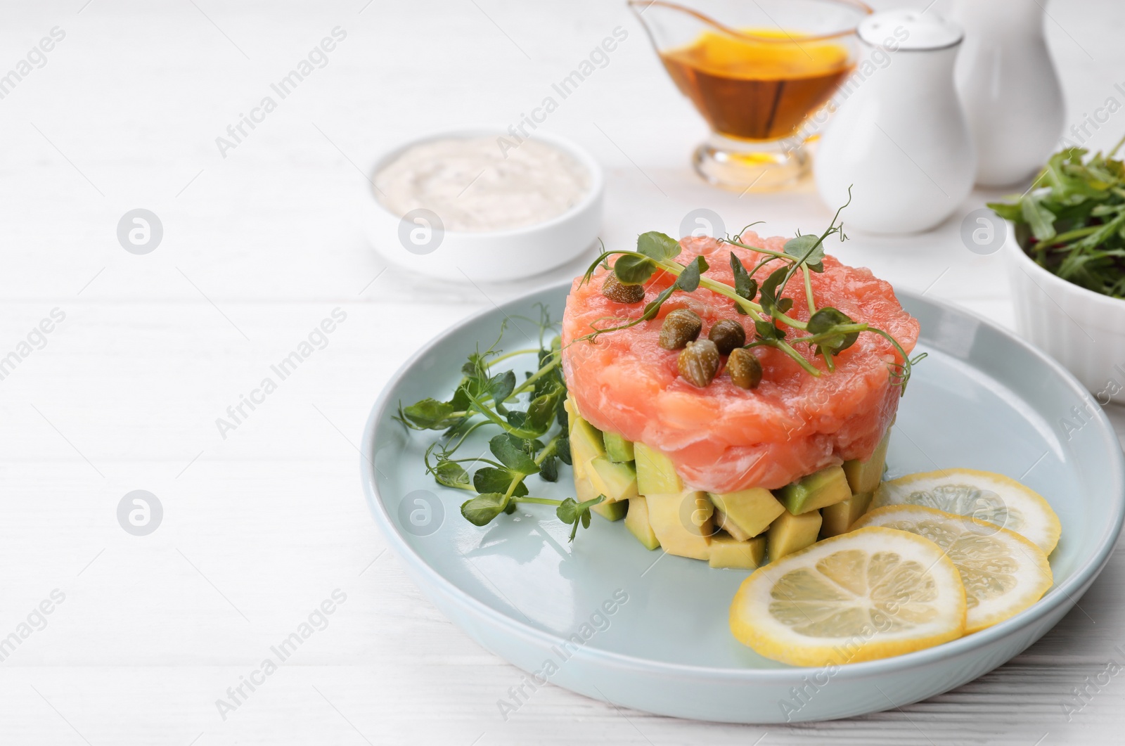 Photo of Tasty salmon tartare with avocado, lemon, capers and microgreens on white wooden table. Space for text