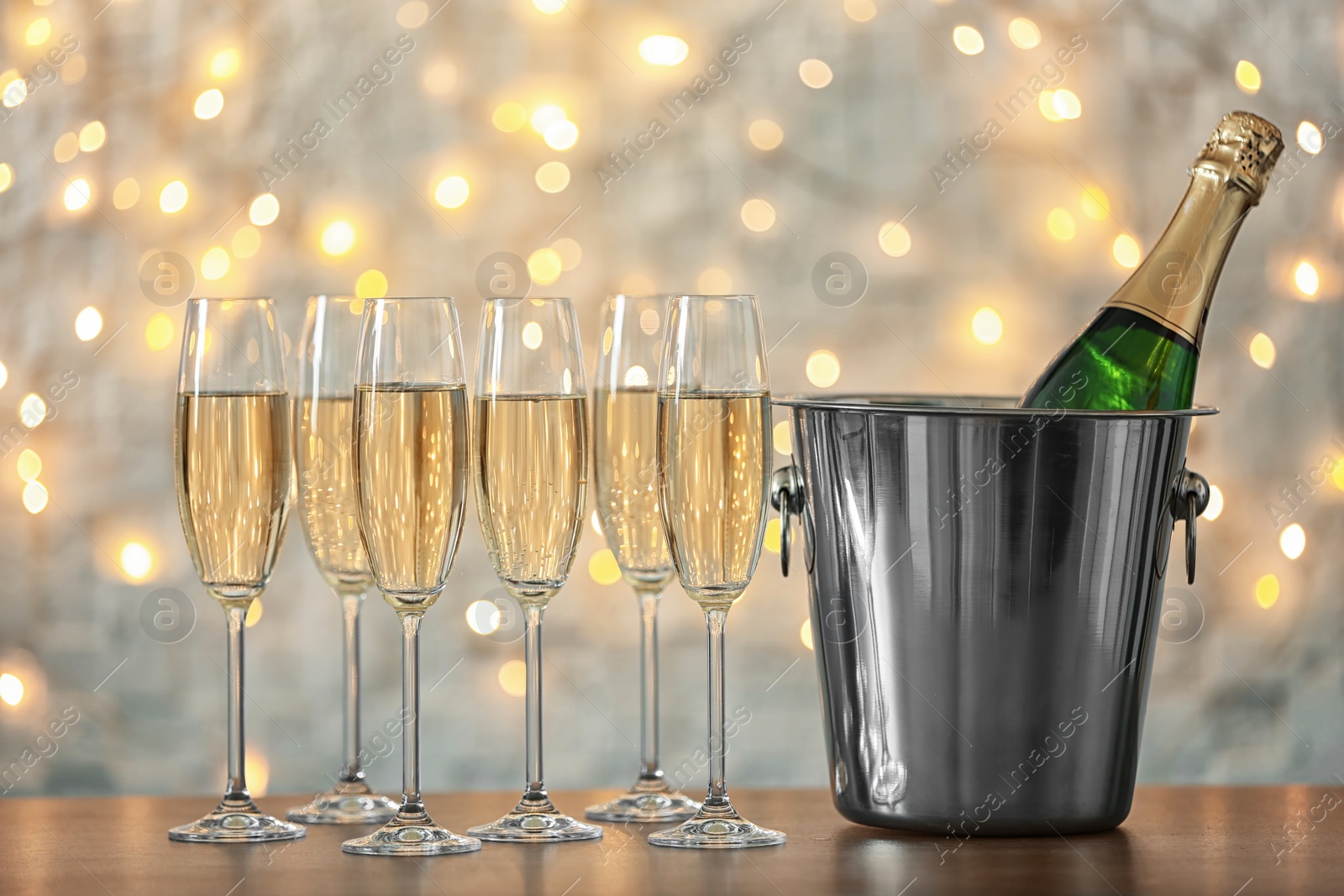 Photo of Glasses with champagne and bottle in bucket on table against blurred lights