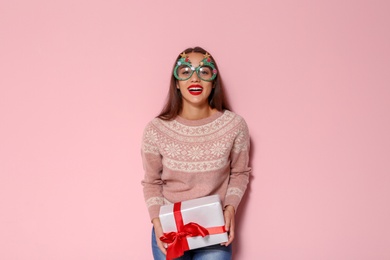 Young woman in warm sweater with Christmas gift and party glasses on color background