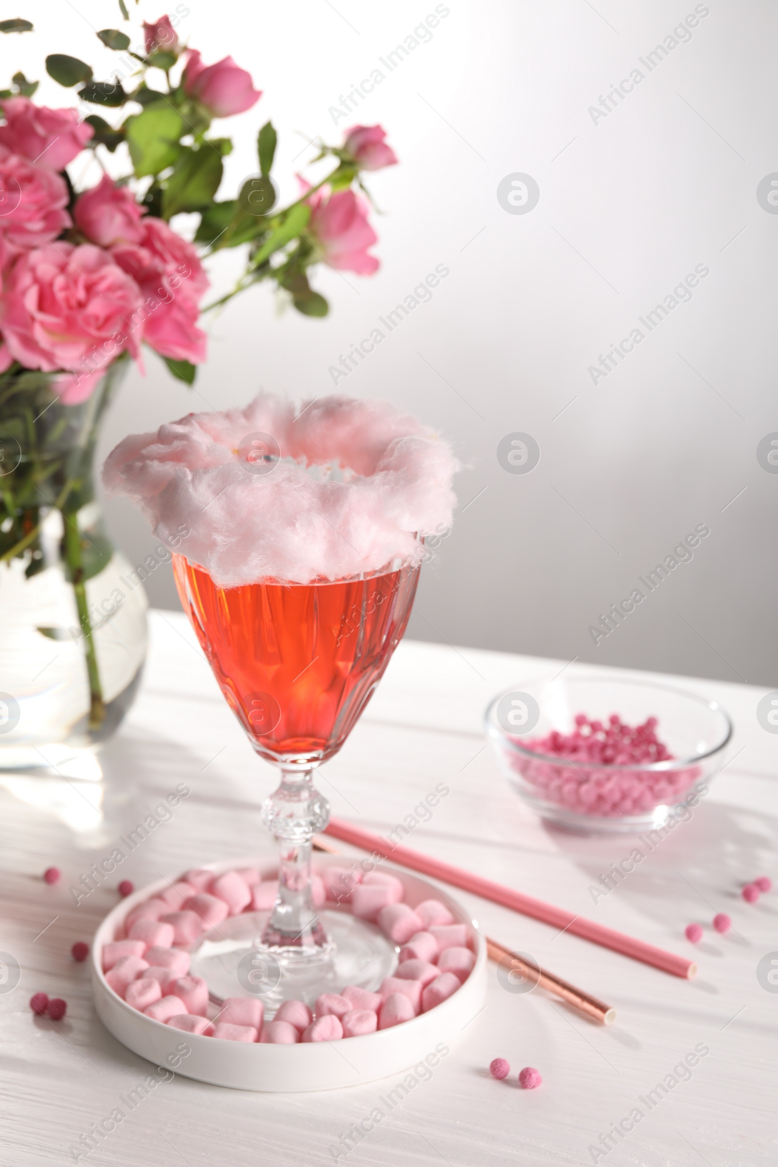 Photo of Cotton candy cocktail in glass, marshmallows, vase with pink roses and straws on white wooden table against gray background