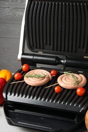 Photo of Electric grill with homemade sausages, rosemary and tomatoes on table