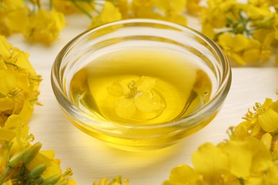 Photo of Rapeseed oil in glass bowl and beautiful yellow flowers on white wooden table, closeup