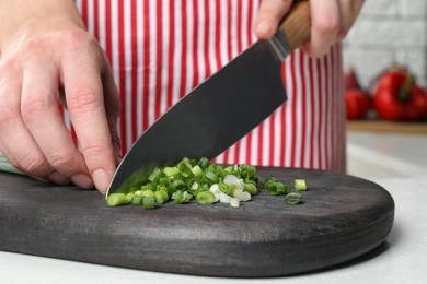 Photo of Woman cutting green spring onion on black wooden board at white table, closeup