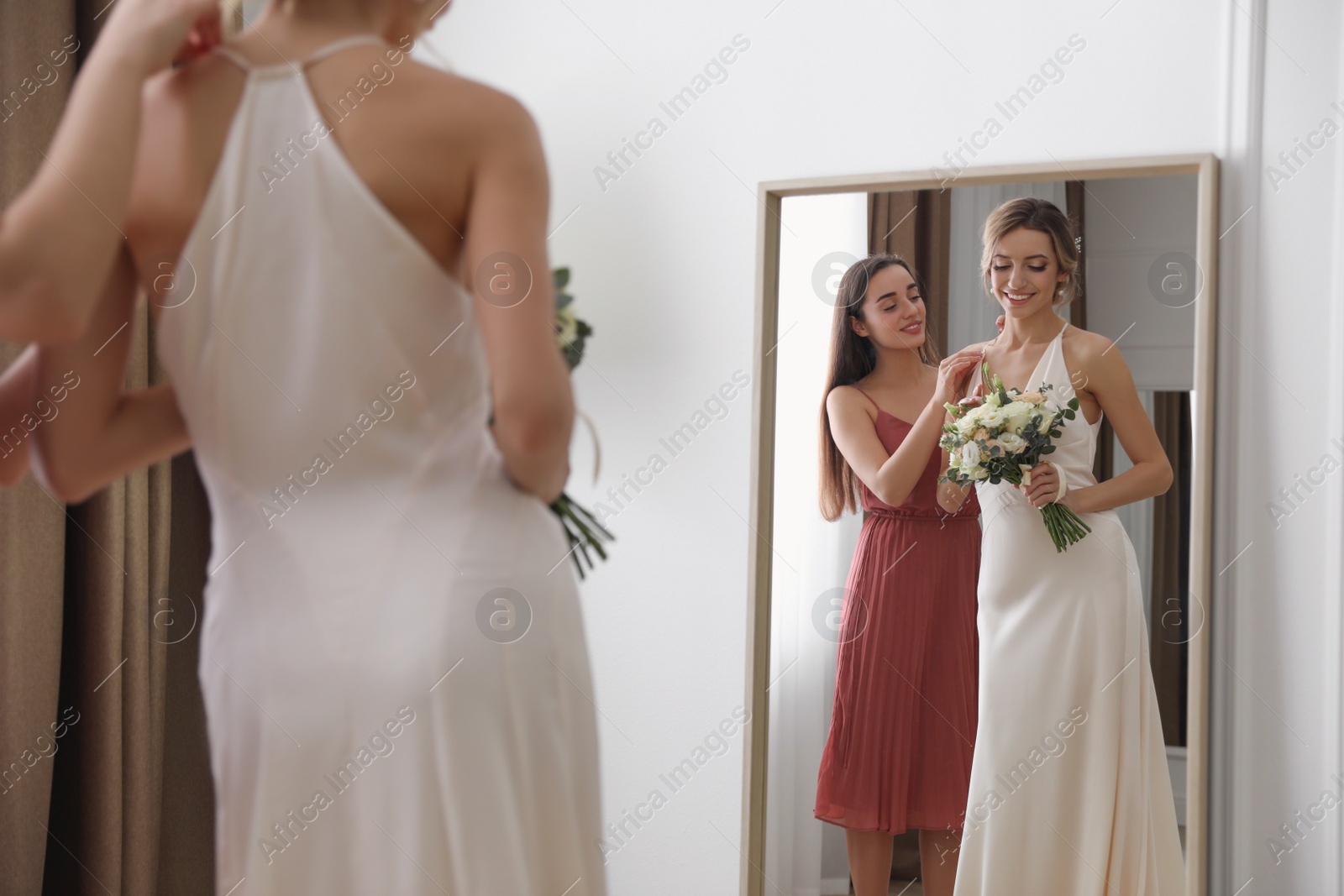 Photo of Gorgeous bride in beautiful wedding dress and her friend near mirror in room