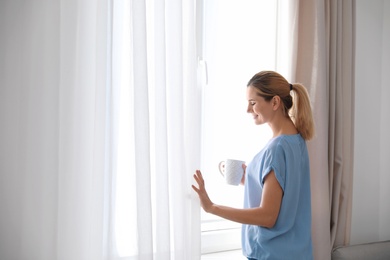 Photo of Woman holding cup of coffee near window with beautiful curtains at home. Space for text