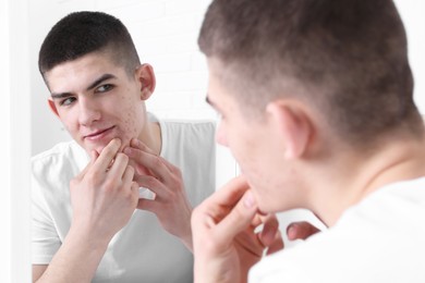 Photo of Young man looking at mirror and touching pimple on his face indoors. Acne problem