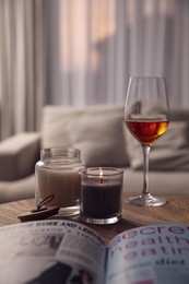 Glass of wine, burning candles and magazine on wooden table in living room