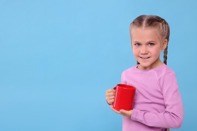 Happy girl with red ceramic mug on light blue background, space for text