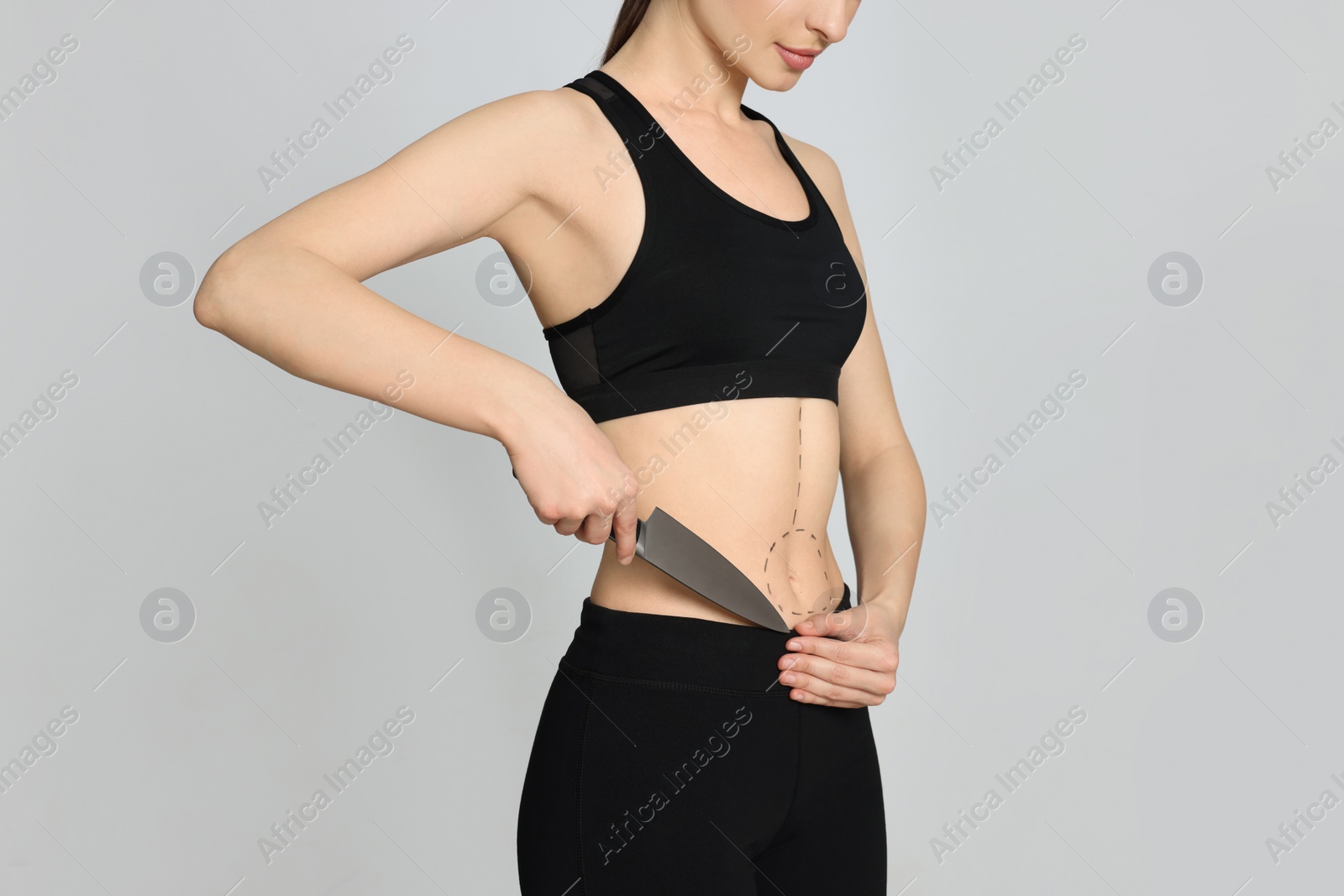 Photo of Slim young woman with knife and marks on body against light background, closeup. Weight loss surgery