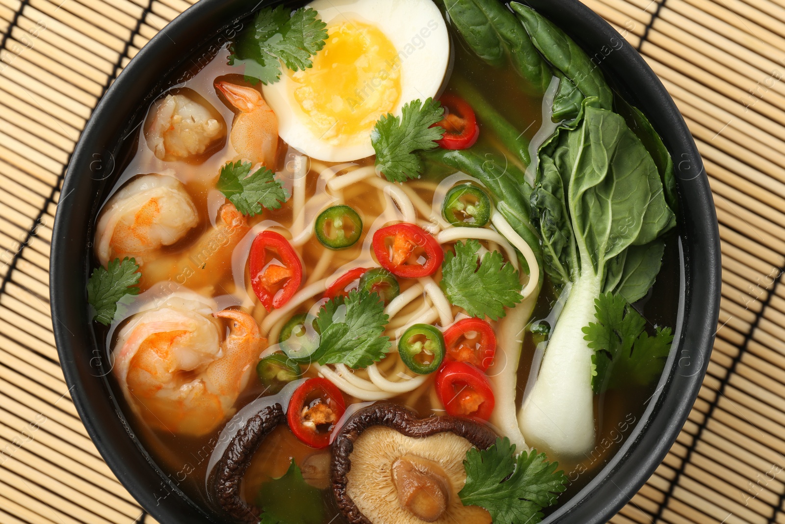 Photo of Delicious ramen with shrimps and egg in bowl on bamboo mat, top view. Noodle soup